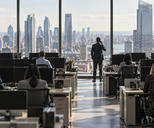 Synthetic editorial illustration. A trading floor. Rear-facing workers sit among rows of desks and dark monitors, while a suited colleague stands by tall windows holding a phone to an ear above a bright midday city view.