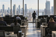 Synthetic editorial illustration. A trading floor. Rear-facing workers sit among rows of desks and dark monitors, while a suited colleague stands by tall windows holding a phone to an ear above a bright midday city view.