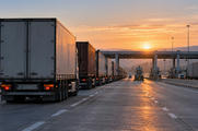 Synthetic editorial illustration. A motorway border checkpoint. A long line of plain freight trailers stretches toward inspection booths beneath a broad canopy, with low dawn sunlight warming the empty adjacent lane.