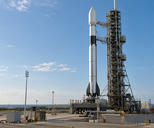 Synthetic editorial illustration. Generic unbranded space-launch vehicle beside a practical access tower on a launch pad; no launch in progress or real named vehicle/site.