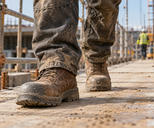 Synthetic editorial illustration. A construction-site walkway. Scuffed brown safety boots and dusty work-trouser legs fill the low foreground, with scaffold rails, stacked timber and a small rear-facing worker blurred behind.