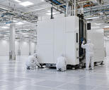 Synthetic editorial illustration. A new semiconductor fabrication bay. Three workers in hooded cleanroom suits seen from behind align a large white enclosed machine and its service connections on an otherwise open raised floor.