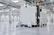 Synthetic editorial illustration. A new semiconductor fabrication bay. Three workers in hooded cleanroom suits seen from behind align a large white enclosed machine and its service connections on an otherwise open raised floor.