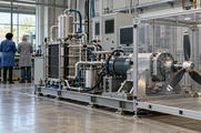 Synthetic editorial illustration. An aerospace test laboratory. A metal stack and pipework connect to an electric motor and stationary propeller inside a clear enclosure; two engineers stand with their backs to the camera by a bench.