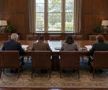 Synthetic editorial illustration. A traditional boardroom. Four trustees seen from behind review papers at a long wooden table, with empty leather chairs opposite and daylight through tall windows.