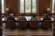 Synthetic editorial illustration. A traditional boardroom. Four trustees seen from behind review papers at a long wooden table, with empty leather chairs opposite and daylight through tall windows.