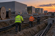 Synthetic editorial illustration. A data-centre construction site. Three workers viewed from behind pull thick power cables along an open trench beside cable reels and unfinished concrete halls at dusk.