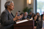Synthetic editorial illustration. An older speaker addresses an audience at a lectern with a mounted microphone and populated notes.