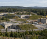 Synthetic editorial illustration. A forested industrial compound. Low concrete buildings sit between grassy earth berms, while a crane rises beside a new concrete structure and pine trees surround the site.