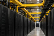 Synthetic editorial illustration. A data-centre hall has yellow fibre ducts and black cable trays overhead. Parallel rows of black cabinets flank an empty aisle leading toward a distant doorway.