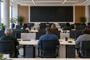 Synthetic editorial illustration. An operations office. Staff seen from behind sit at rows of desks with monitor backs facing the camera and a large dark wall display ahead, in a bright windowed room.