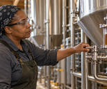 Synthetic editorial illustration. A brewery worker holds a valve handle beside a liquid-filled sight glass on connected tank pipework.