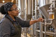 Synthetic editorial illustration. A brewery worker holds a valve handle beside a liquid-filled sight glass on connected tank pipework.