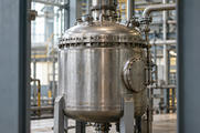 Synthetic editorial illustration. One sealed stainless industrial process vessel, inspection hatch and pipe connections in an anonymous processing hall, framed between structural supports.