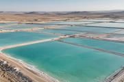 Synthetic editorial illustration. Broad turquoise evaporation ponds are divided by narrow pale earth berms across an arid plain. A low aerial view reveals varied blue-green water and distant barren hills under hazy daylight.
