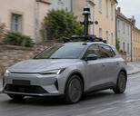 Synthetic editorial illustration. A plain grey car carries a tall roof-mounted mast with cameras and a cylindrical sensor through a quiet town street. Modest motion blur runs past unmarked plaster facades and a stone wall under overcast light.