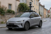 Synthetic editorial illustration. A plain grey car carries a tall roof-mounted mast with cameras and a cylindrical sensor through a quiet town street. Modest motion blur runs past unmarked plaster facades and a stone wall under overcast light.
