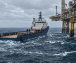 Synthetic editorial illustration. An offshore work site. A blue supply vessel with an open aft deck sits alongside a steel platform above choppy waves and whitecaps under an overcast sky.