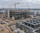 Synthetic editorial illustration. A data-centre construction site contains steel-framed buildings and tower cranes. Cooling units, exposed pipes and stacked materials fill the broad site beneath an overcast sky.