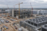 Synthetic editorial illustration. A data-centre construction site contains steel-framed buildings and tower cranes. Cooling units, exposed pipes and stacked materials fill the broad site beneath an overcast sky.