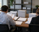 Synthetic editorial illustration. Two people viewed from behind review blank sheets in open binders at a wooden office table. Stacks of plain white binders and shelves of unmarked files surround them in soft daylight.