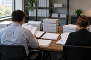 Synthetic editorial illustration. Two people viewed from behind review blank sheets in open binders at a wooden office table. Stacks of plain white binders and shelves of unmarked files surround them in soft daylight.