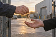 Synthetic editorial illustration. Two cropped adults exchange a small key ring at a plain industrial-property entrance.