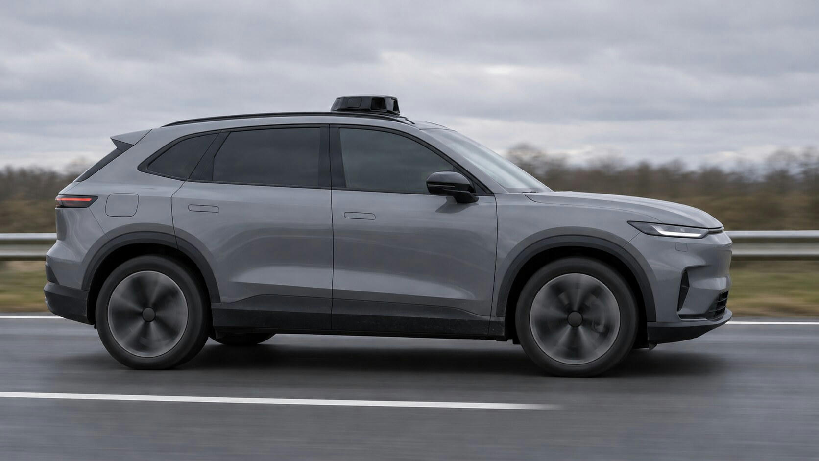 Synthetic editorial illustration. A grey car with dark windows and a small roof sensor pod travels along a motorway. The side-on view shows spinning wheels, a blurred guardrail and trees beneath an overcast sky.