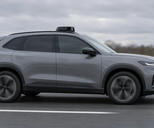 Synthetic editorial illustration. A grey car with dark windows and a small roof sensor pod travels along a motorway. The side-on view shows spinning wheels, a blurred guardrail and trees beneath an overcast sky.
