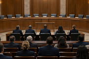 Synthetic editorial illustration. A legislative hearing room. Witnesses and attendees seen from behind face a raised curved dais with empty chairs, blank nameplates and microphones against wood-panelled walls.