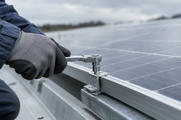 Synthetic editorial illustration. A gloved hand uses a ratchet on a metal mounting clamp at the edge of a photovoltaic panel, with the array softly receding behind.