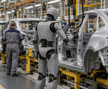 Synthetic editorial illustration. A car assembly line. A grey articulated humanoid robot stands beside a bare metal vehicle shell and suspended tools, while two rear-facing workers in plain coveralls walk along the adjacent aisle.