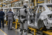 Synthetic editorial illustration. A car assembly line. A grey articulated humanoid robot stands beside a bare metal vehicle shell and suspended tools, while two rear-facing workers in plain coveralls walk along the adjacent aisle.
