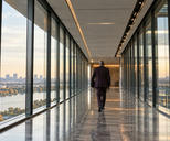 Synthetic editorial illustration. A glass office corridor overlooks water and a distant city. A rear-facing suited person carrying a dark folder walks away along the reflective stone floor.