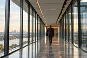 Synthetic editorial illustration. A glass office corridor overlooks water and a distant city. A rear-facing suited person carrying a dark folder walks away along the reflective stone floor.
