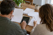 Synthetic editorial illustration. Two people viewed from behind handle blank papers and envelopes at a wooden kitchen table. One uses a calculator with unmarked keys and a dark display beside a plain mug and a potted plant.
