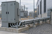 Synthetic editorial illustration. A deployed electrical cabinet feeds multiple supported conduit runs into a facility wall, with switchyard equipment behind.