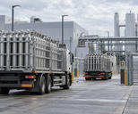 Synthetic editorial illustration. A fabrication-plant utility yard. Two trucks carry cages of upright metal gas cylinders through an open gate, with storage tanks and pipe bridges behind them under cloudy skies.