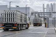 Synthetic editorial illustration. A fabrication-plant utility yard. Two trucks carry cages of upright metal gas cylinders through an open gate, with storage tanks and pipe bridges behind them under cloudy skies.