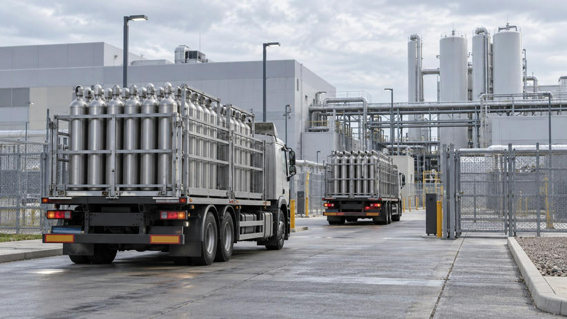Synthetic editorial illustration. A fabrication-plant utility yard. Two trucks carry cages of upright metal gas cylinders through an open gate, with storage tanks and pipe bridges behind them under cloudy skies.