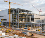 Synthetic editorial illustration. A large industrial building under construction shows exposed steel framing, pipes and partly installed white cladding. Tower cranes, small construction vehicles and tiny workers occupy the orderly site under pale daylight.