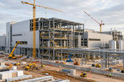 Synthetic editorial illustration. A large industrial building under construction shows exposed steel framing, pipes and partly installed white cladding. Tower cranes, small construction vehicles and tiny workers occupy the orderly site under pale daylight.
