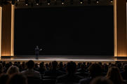 Synthetic editorial illustration. A small presenter viewed from behind gestures on a broad stage before a completely black screen. Rows of audience heads fill the foreground under restrained warm auditorium lighting.