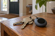 Synthetic editorial illustration. A home entrance. Plain house keys and a rounded dark keychain gadget with a circular black screen rest on a worn wooden table beside a plant pot.