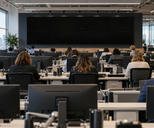 Synthetic editorial illustration. Many people viewed from behind work at rows of newsroom desks beneath ceiling lights. Plain monitor backs fill the foreground and a large bank of completely black screens spans the far wall.