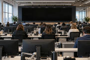 Synthetic editorial illustration. Many people viewed from behind work at rows of newsroom desks beneath ceiling lights. Plain monitor backs fill the foreground and a large bank of completely black screens spans the far wall.