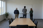 Synthetic editorial illustration. An institutional meeting room. Three colleagues viewed from behind face a whiteboard bearing only coloured curved lines and arrows, beyond a plain wooden table in daylight.