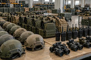 Synthetic editorial illustration. A military equipment depot. Long tables hold rows of helmets, protective vests, handheld radios and night-vision housings, with storage cases and shelving behind them.