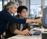 Synthetic editorial illustration. Two developers examine a visible populated desktop monitor, one pointing while the seated colleague uses a mouse.