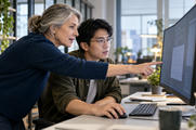 Synthetic editorial illustration. Two developers examine a visible populated desktop monitor, one pointing while the seated colleague uses a mouse.